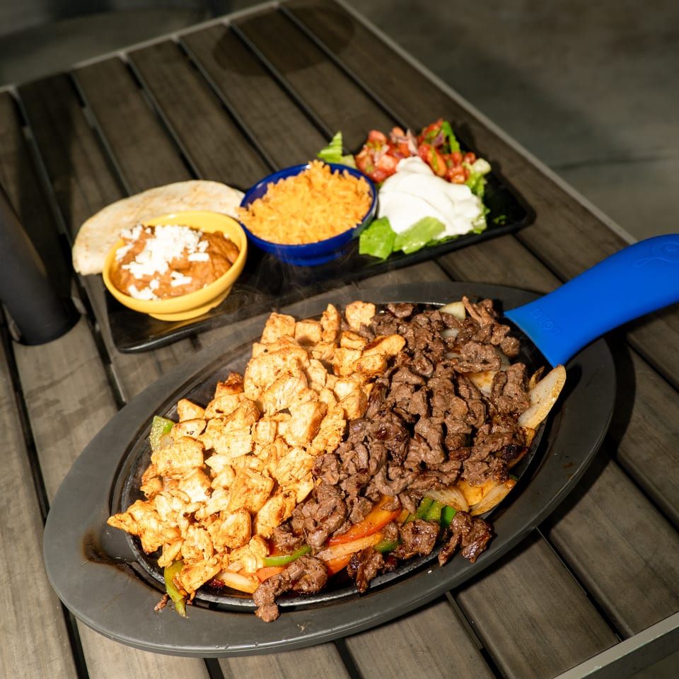 Sizzling chicken and steak fajitas with sides of rice, beans, tortillas, and salad on a wooden table.