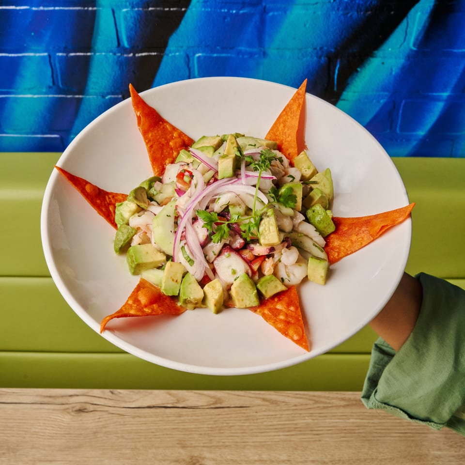 A person holds a white plate of ceviche with avocado, red onion, and tortilla chips.