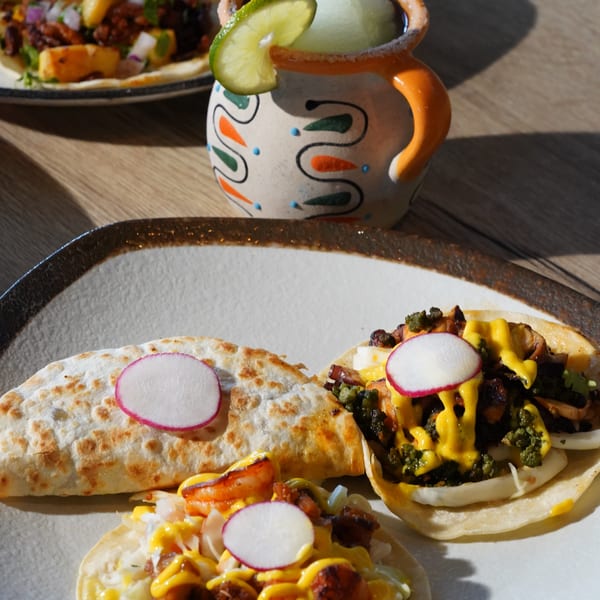 A plate of tacos and a quesadilla with radishes, alongside a frozen margarita in a colorful mug.