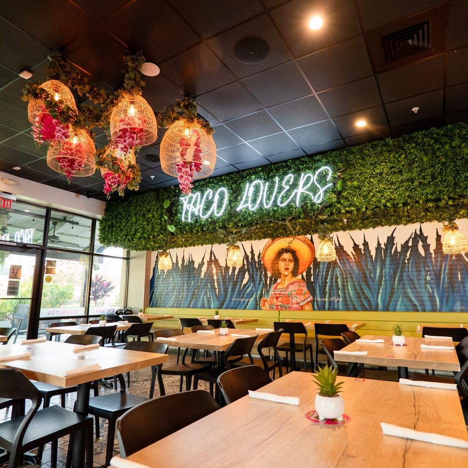 Restaurant interior with wooden tables, black chairs, and a neon "TACO LOVERS" sign above a mural.