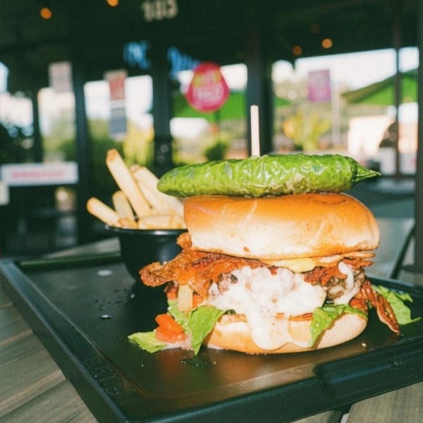 Gourmet burger with melted cheese, bacon, lettuce, tomato, and a whole pickle on top, with fries.
