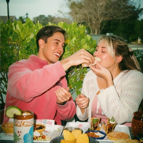 A smiling man feeds a taco to a woman at an outdoor restaurant table filled with Mexican food.